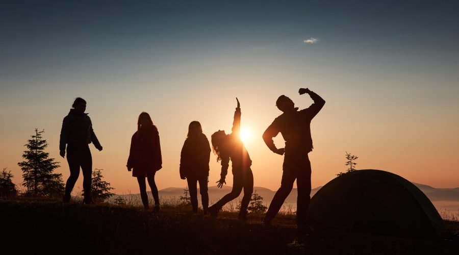 A silhouette of group people have fun at the top of the mountain near the tent during the sunset.
